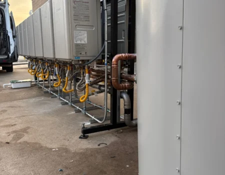 Commercial tankless water heater system installed outdoors, featuring multiple units connected to a large storage tank with copper piping and gas lines.